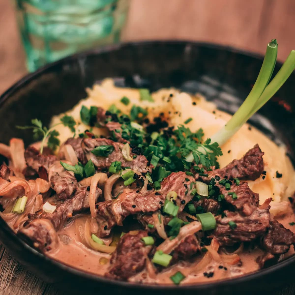 Beef stroganoff with soft onions in a creamy sauce, topped with spring onions and parsley, on mashed potato in a dark bowl