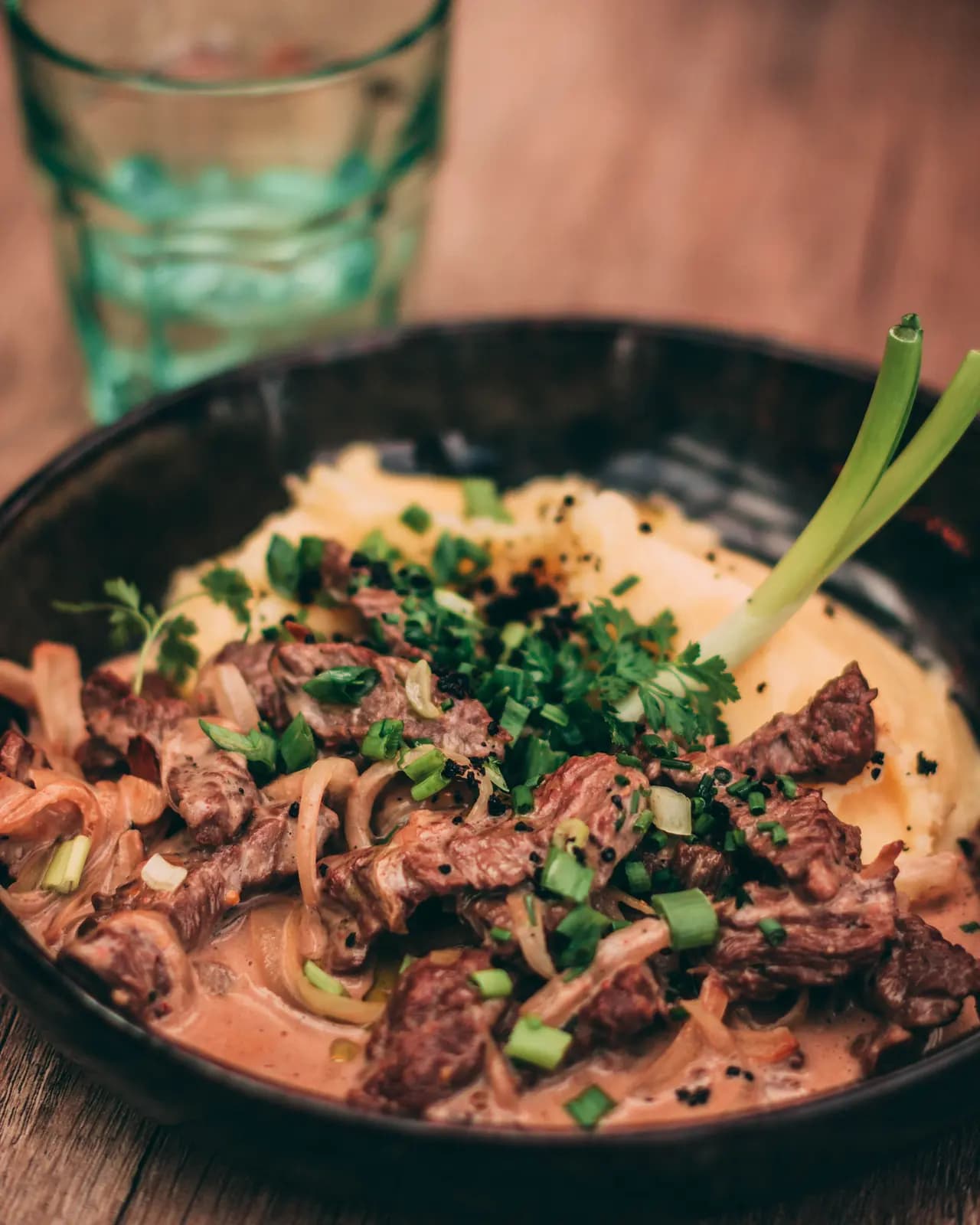 Beef stroganoff with soft onions in a creamy sauce, topped with spring onions and parsley, on mashed potato in a dark bowl