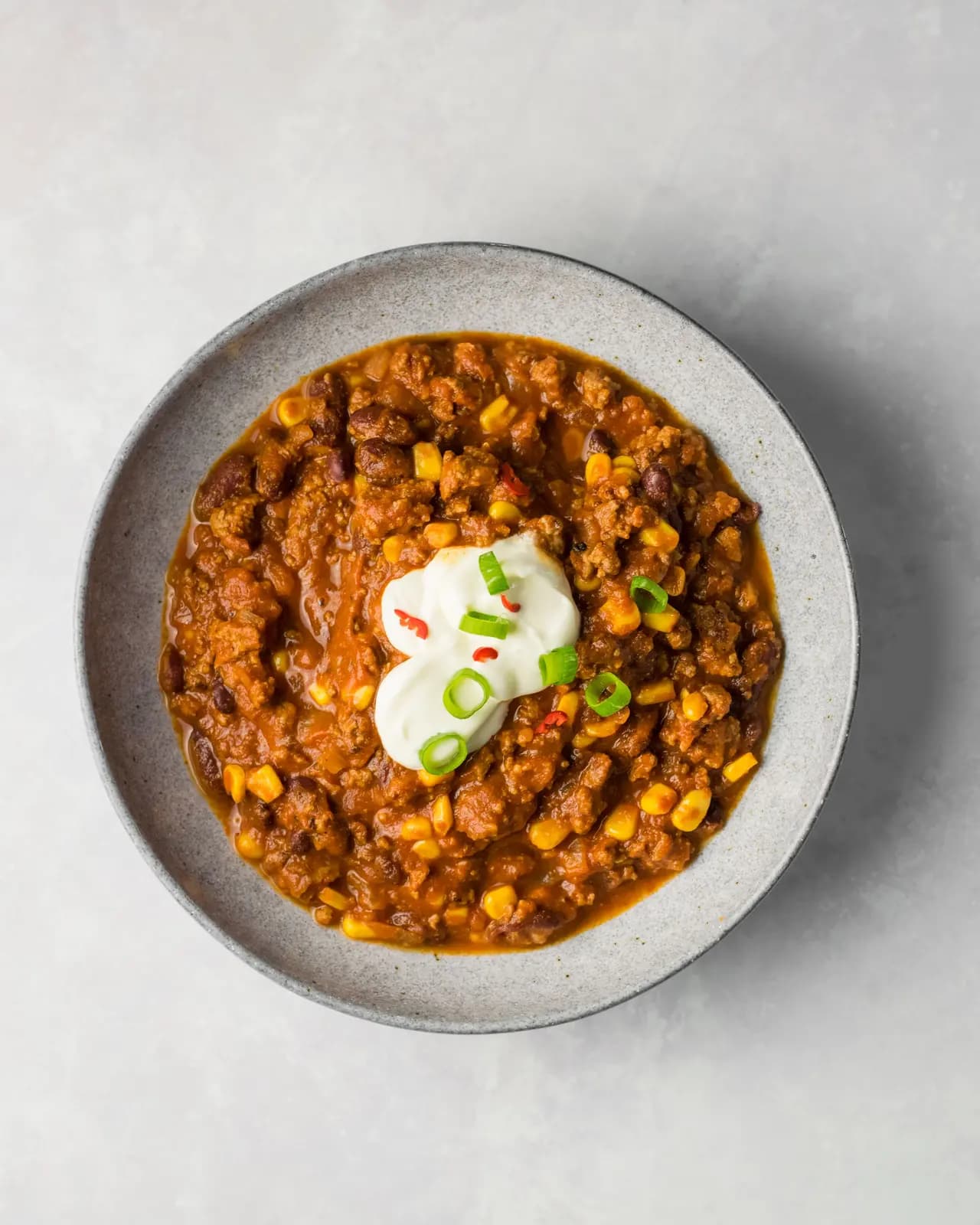Beef chili with kidney beans and corn, topped with sour cream, green onions and red chili in a gray bowl