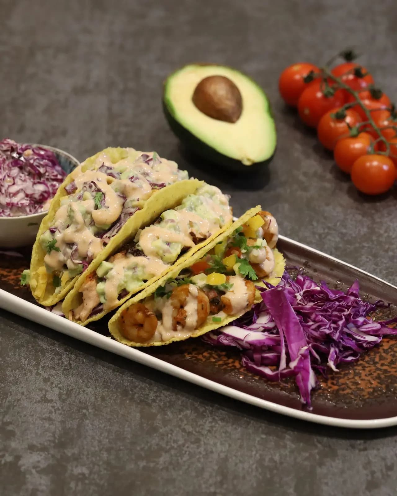 Three prawn tacos in crisp shells with red cabbage slaw, avocado and pink sauce, beside an avocado half and cherry tomatoes