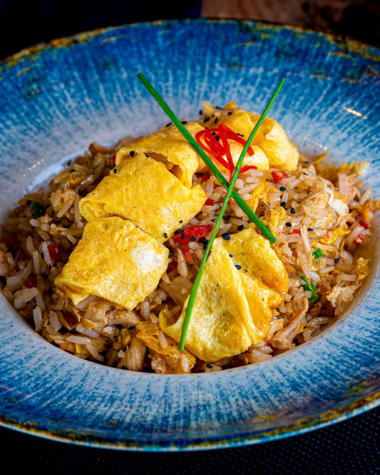 Egg fried rice with red pepper, a folded omelette, sliced red chilli, chives and black sesame seeds in a blue bowl