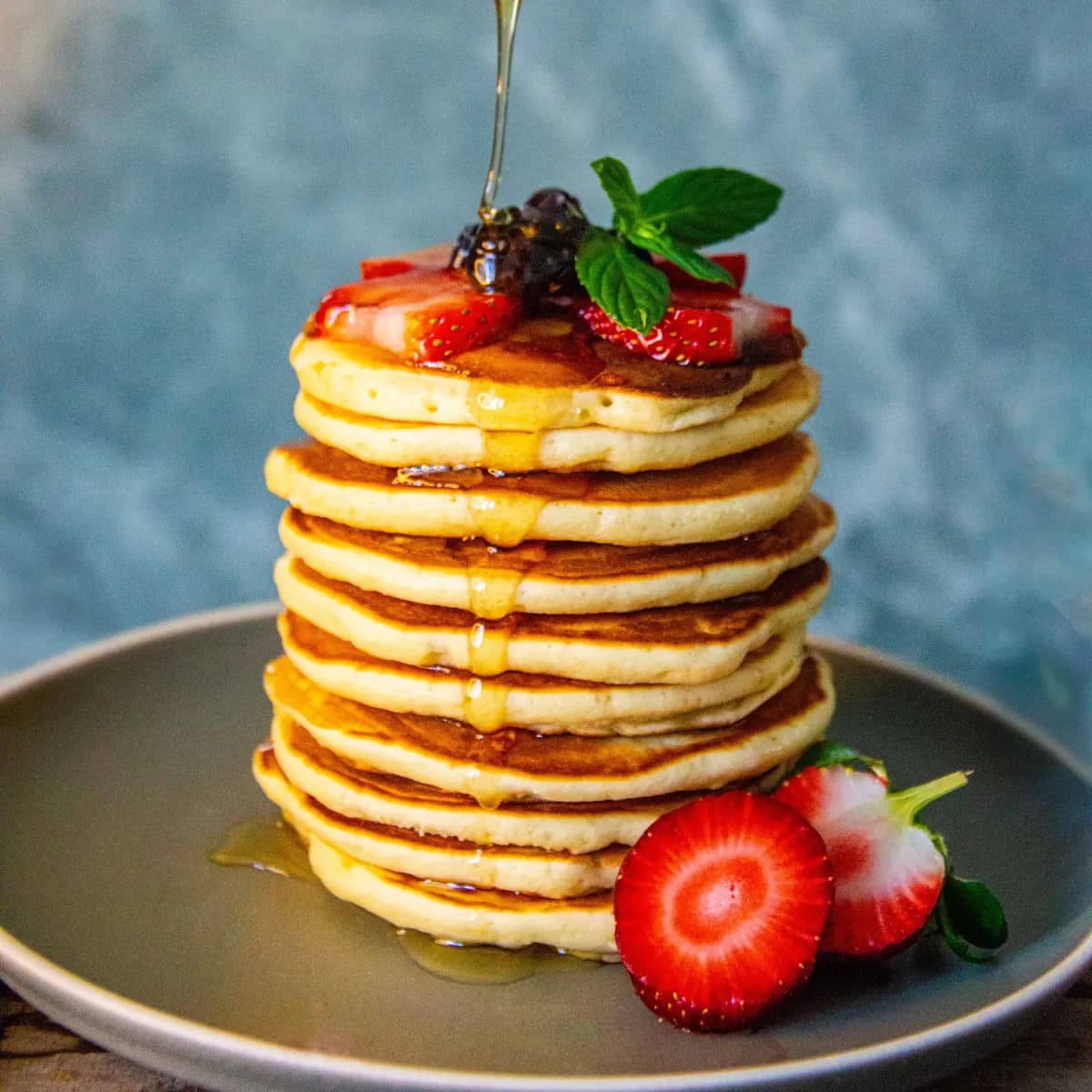 A tall stack of fluffy pancakes topped with strawberries, blackberries and mint, with honey drizzling from a wooden dipper