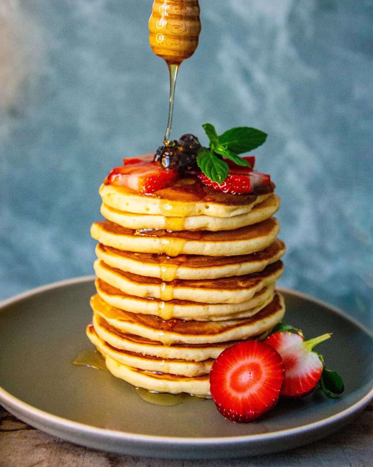 A tall stack of fluffy pancakes topped with strawberries, blackberries and mint, with honey drizzling from a wooden dipper