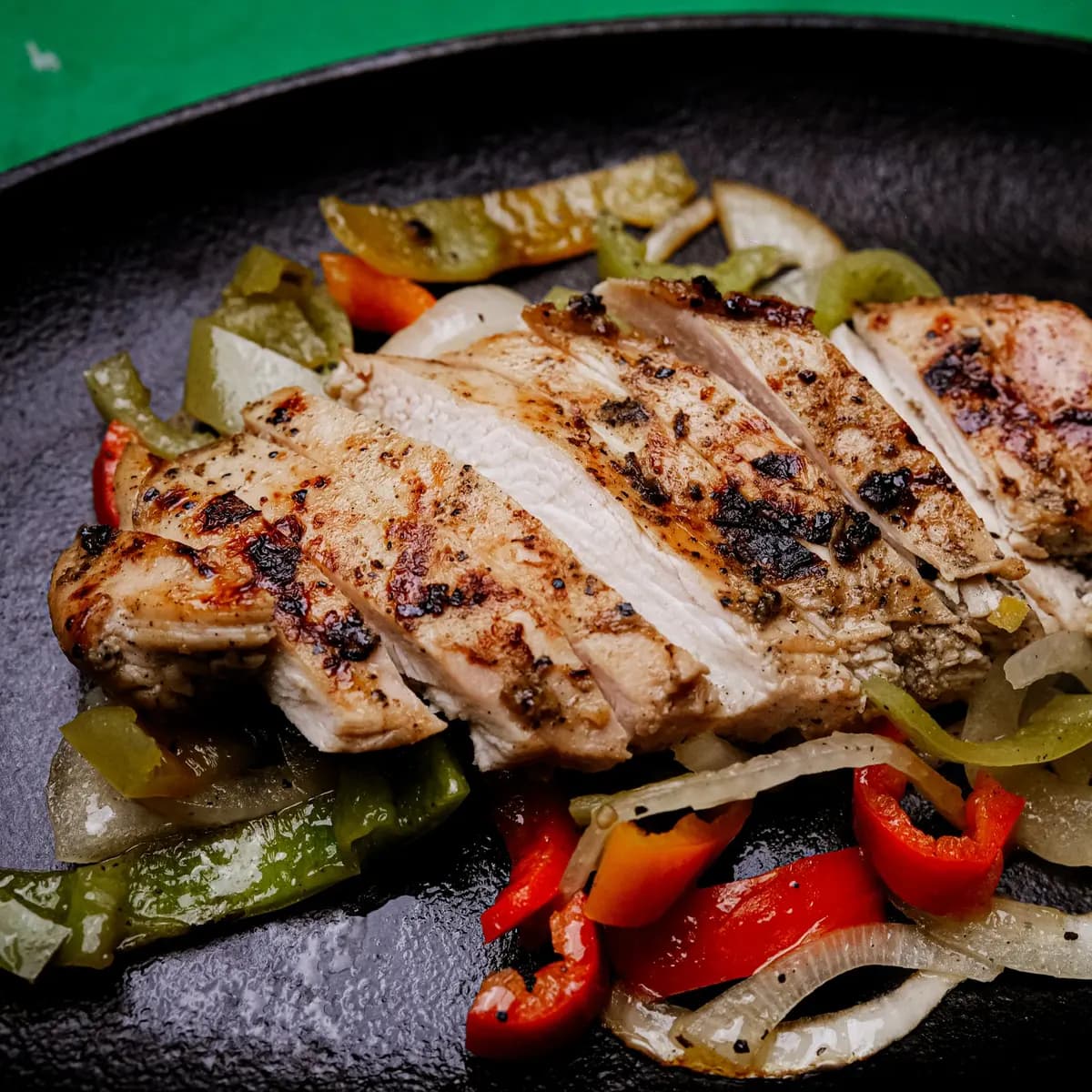 Sliced spiced chicken breast on strips of red and green pepper and onion on a black cast iron plate