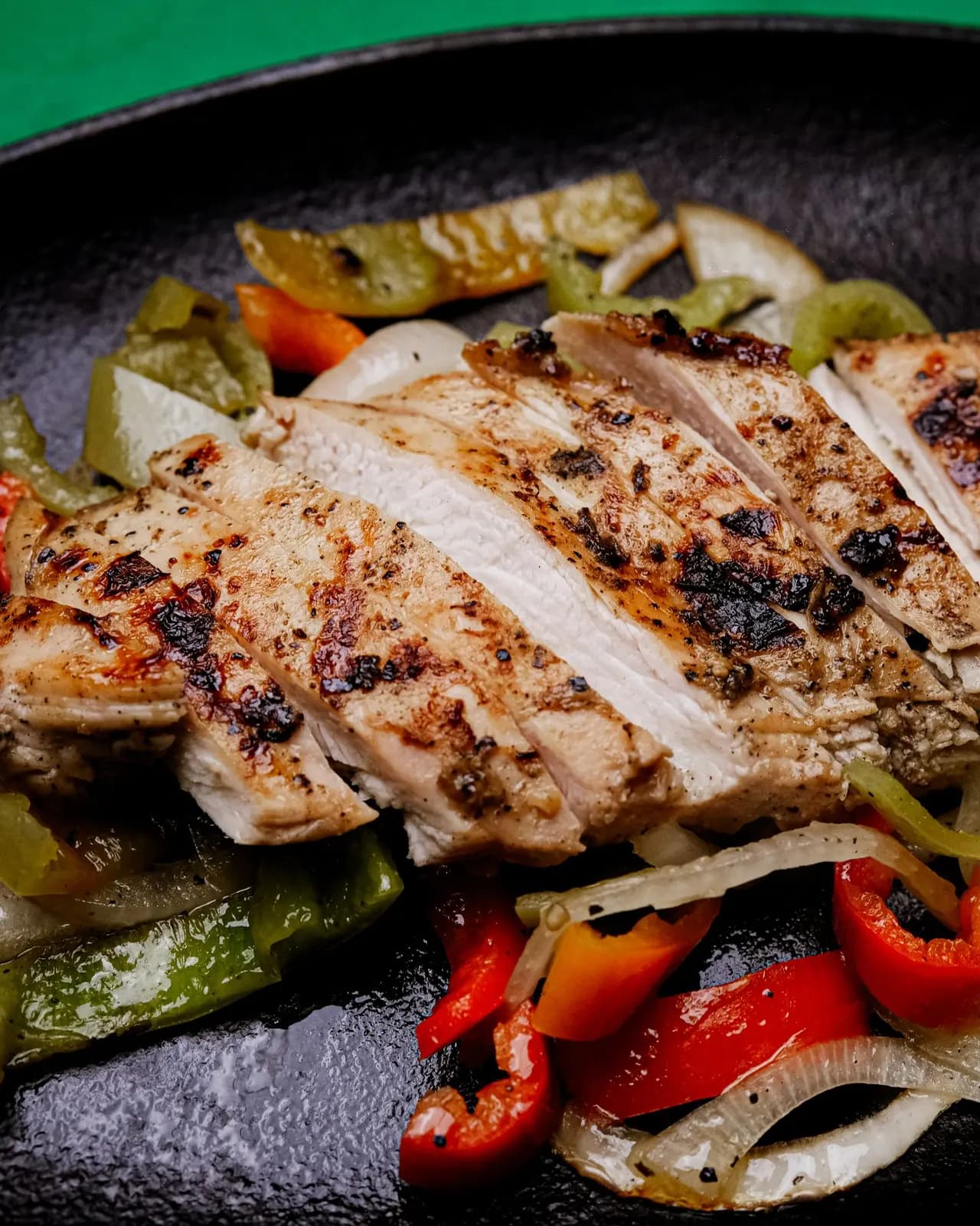 Sliced spiced chicken breast on strips of red and green pepper and onion on a black cast iron plate