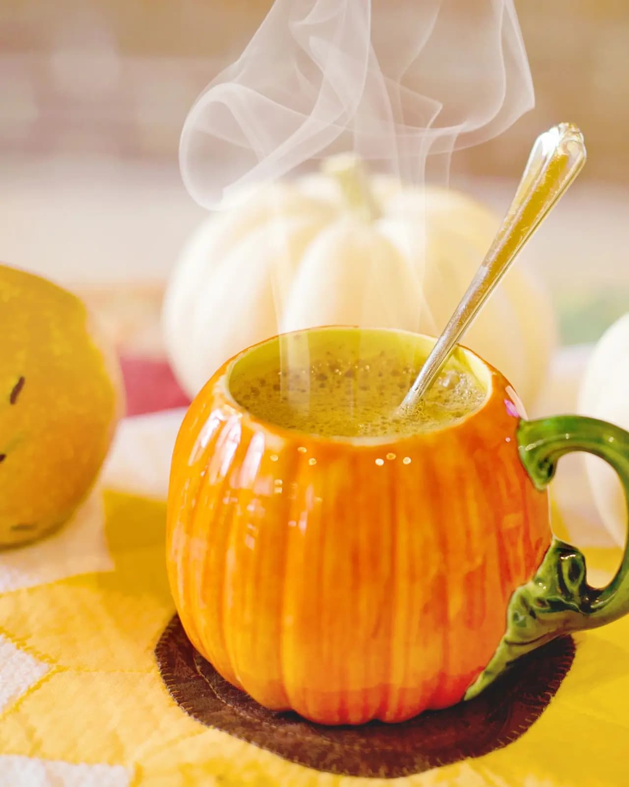 Pumpkin spice latte with a speckled froth in an orange pumpkin shaped mug with a spoon, steam rising, beside two white pumpkins