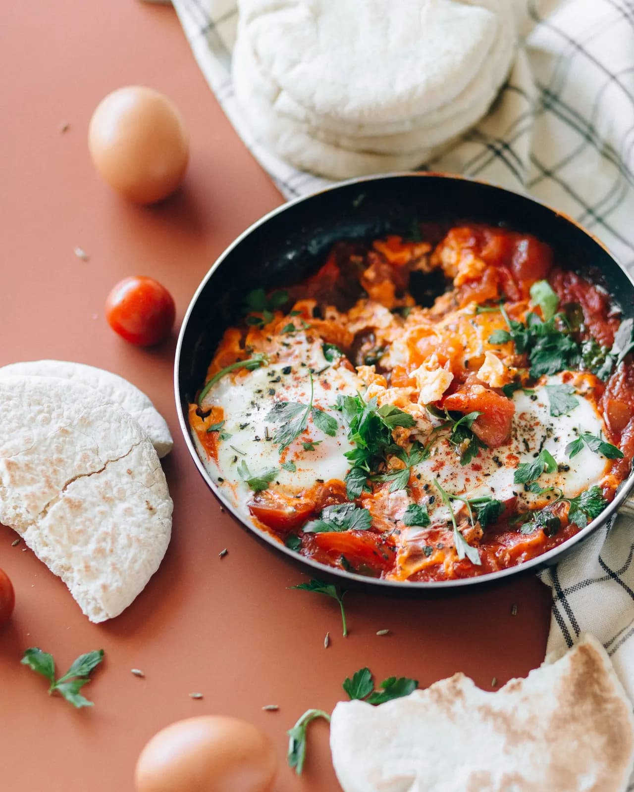 Shakshuka with eggs, chunks of tomato and parsley in a small black frying pan, with pitta breads, eggs and cherry tomatoes around it