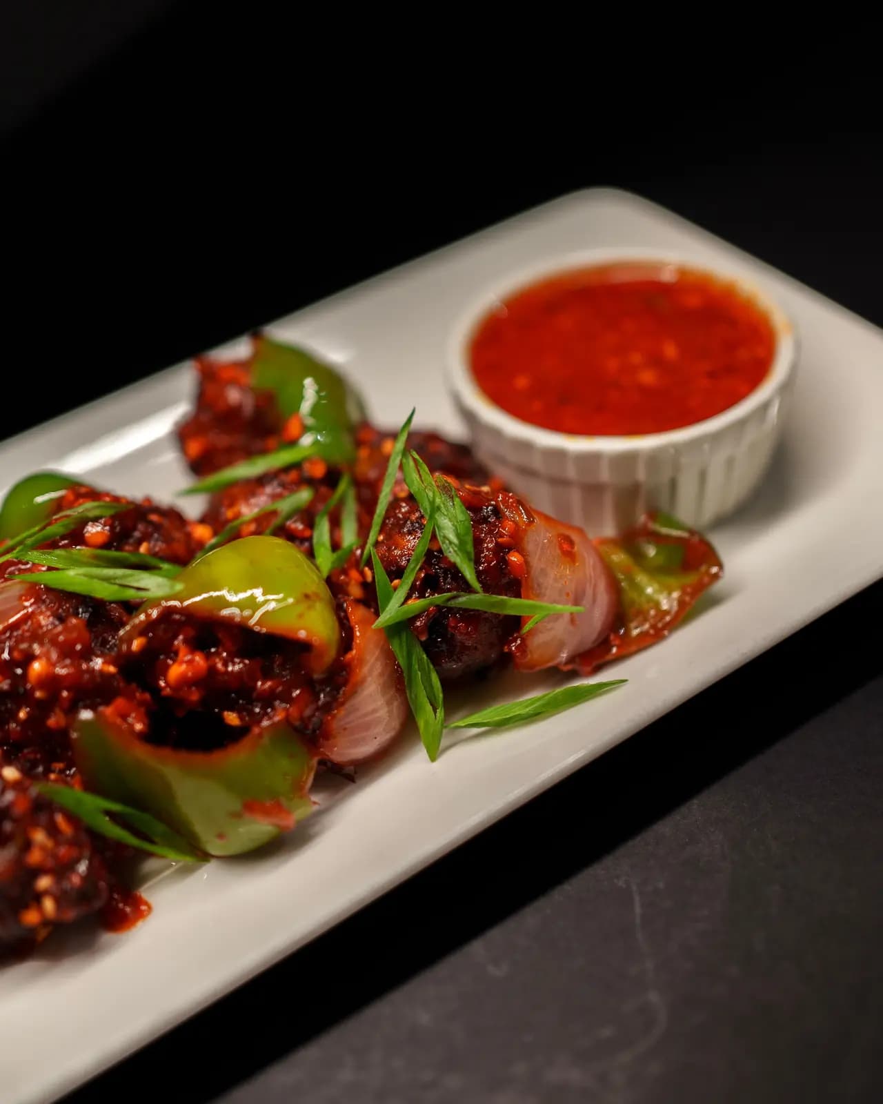 Chicken pieces with chilli flakes, green pepper, red onion and spring onion on a white plate, with a pot of chilli dipping sauce