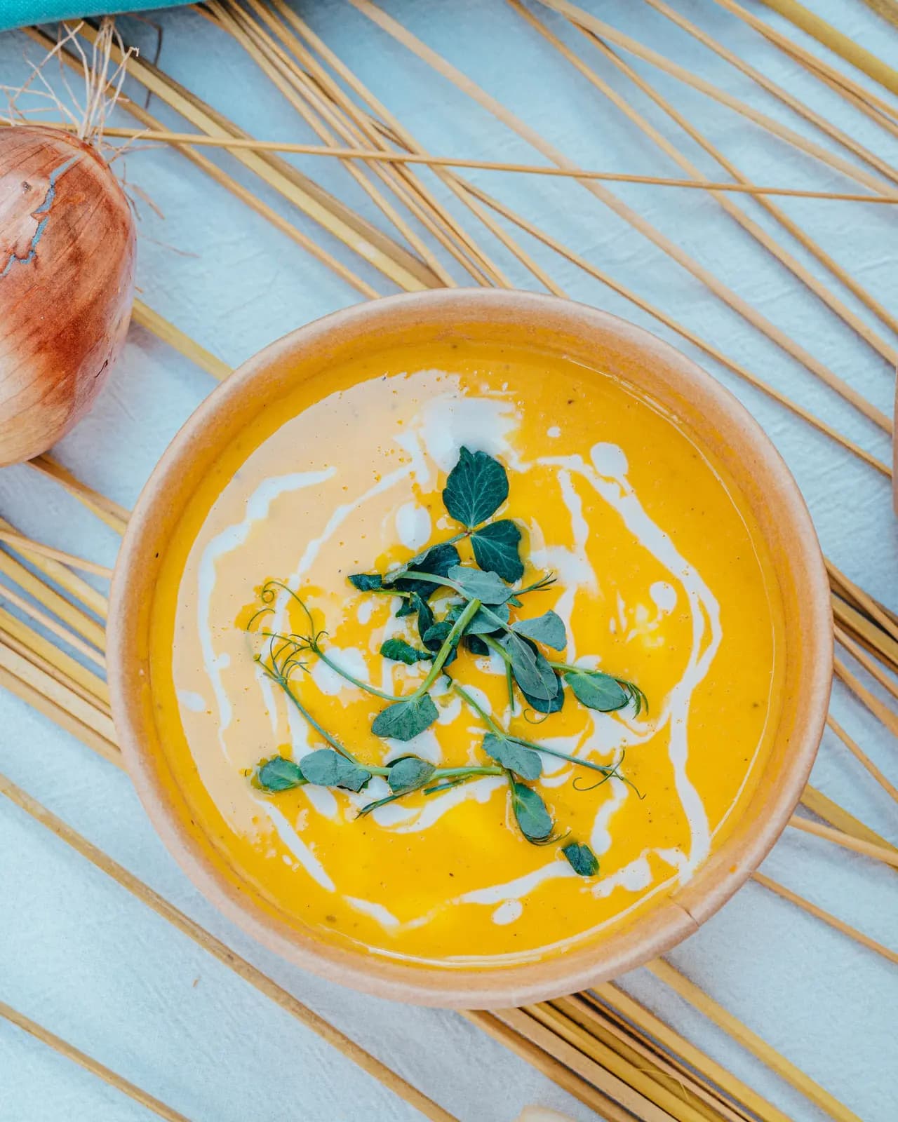 Smooth orange pumpkin soup with a swirl of cream and pea shoots in a paper bowl, beside an onion